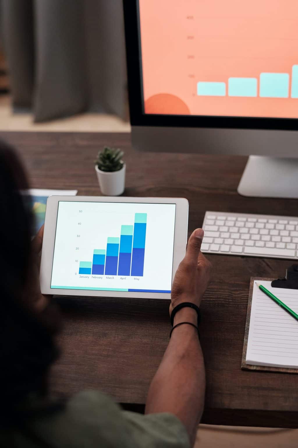 Person analysing sales growth chart on tablet with computer monitor in background.